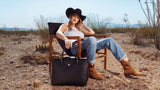 Model seated outdoors in a desert landscape with the Alessia Large Tote in black ostrich beside her