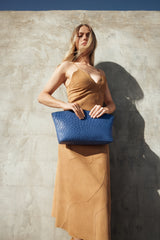 Model standing against a textured wall holding the Blake Large Clutch in blue ostrich