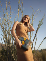 Model standing among desert grasses under a blue sky with the Stevie Medium Clutch in desert sky python