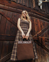 Model in a plaid dress standing in front of a rustic wooden wall with the Gisele Large Shopper in brown suede