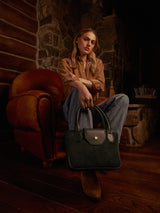 Model seated in a leather armchair in a warmly lit room with the Lucille Mini Tote in bottle green suede